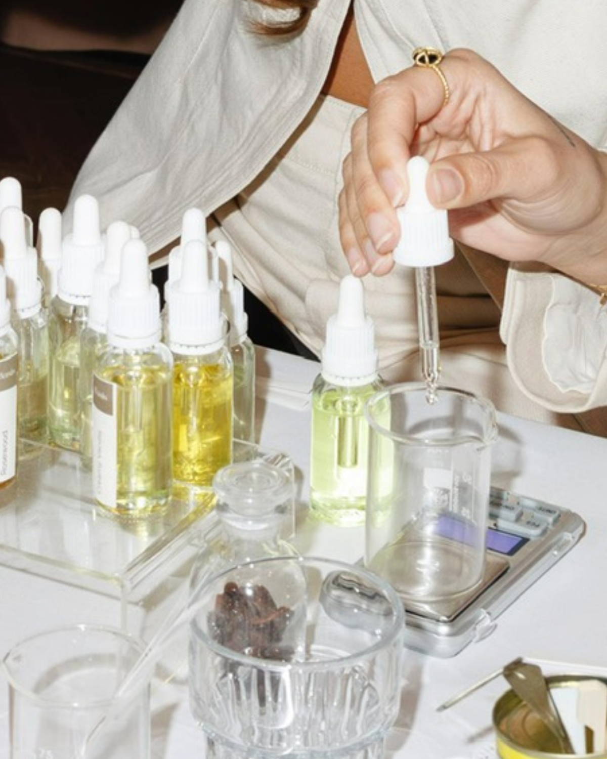 A close-up of a person's hands holding a white dropper over a glass beaker on a scale, with multiple small dropper bottles containing yellow and green liquids arranged on a table.